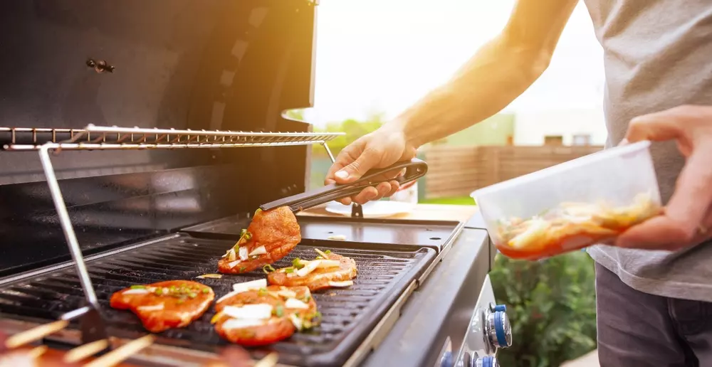 Churrasqueira a gás com carne sendo temperada e grelhada por pessoa em ambiente externo, destacando a praticidade e o sabor na preparação de alimentos.