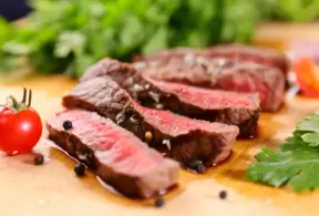 Carne vermelha ao ponto, destacando a presença de mioglobina que dá a cor rosa e vermelha à carne, com tomate e folhas verdes ao fundo.