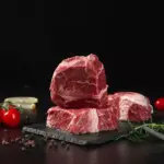 Três pedaços de carne vermelha com tomate cereja, alho e ervas ao redor, destacando a importância dos aminoácidos essenciais na alimentação.