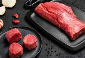 Bife cru de filé mignon — carne vermelha magra — sobre uma tábua de corte preta, com três pedaços de filé mignon em um prato preto, rodeados por alho, cebola e grãos de pimenta