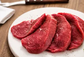 Prato branco com fatias de carne vermelha crua em rodelas grossas, com marcas de gordura e textura, sobre mesa de madeira, Lembrando que ferro heme pode ser encontrado em carne bovina.