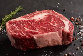 Nem toda gordura da carne é igual: entenda a gordura intramuscular da carne bovina, em um corte de carne vermelha com finas veias de gordura branca sob temperos e sal grosso sobre superfície escura.