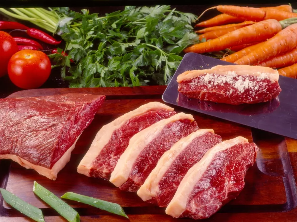 Cortes de carne bovina fresca com vegetais ao fundo, incluindo cenouras, tomate, pimentões e salsa, destacando a qualidade para exportações de carne bovina