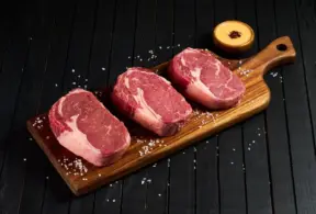 Três cortes de carne vermelha em uma tábua de madeira, simbolizando a importância de padrões dietéticos saudáveis na alimentação, com impacto na nutrição.