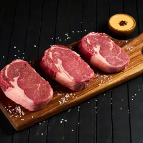 Três cortes de carne vermelha em uma tábua de madeira, simbolizando a importância de padrões dietéticos saudáveis na alimentação, com impacto na nutrição.