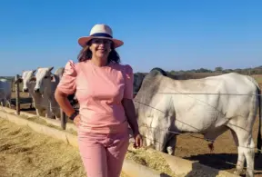 Luzanir Luíza de Moura Peixoto ao ar livre com vacas no fundo, usando chapéu e roupa rosa, em um ambiente rural ensolarado.