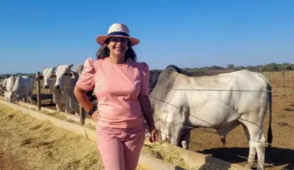 Luzanir Luíza de Moura Peixoto ao ar livre com vacas no fundo, usando chapéu e roupa rosa, em um ambiente rural ensolarado.