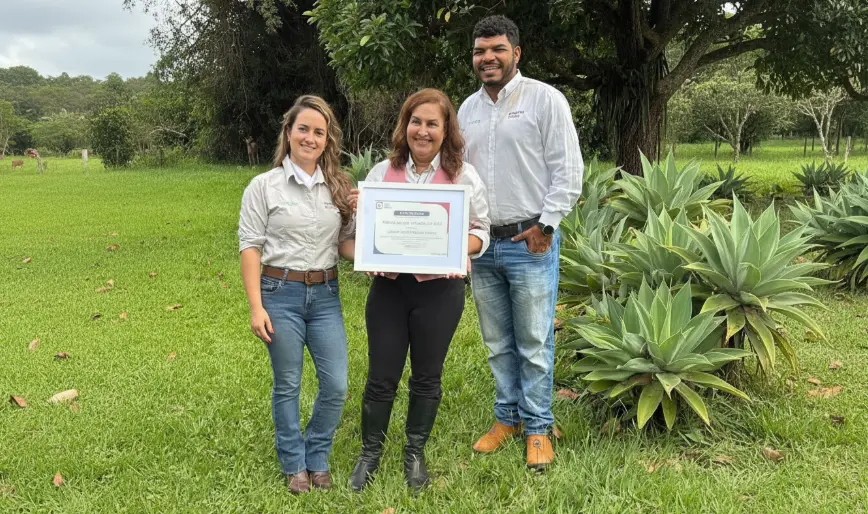Luzanir Luíza de Moura Peixoto recebendo um certificado ao lado de duas pessoas em ambiente ao ar livre, com árvores e plantas ao fundo.