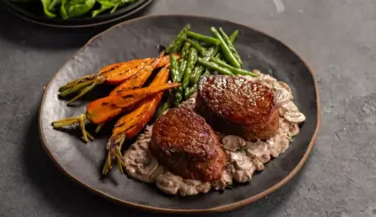 Prato com dois medalhões de carne vermelha ao ponto, acompanhados de legumes assados e molho cremoso