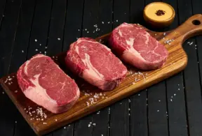Três pedaços de carne bovina fresca, cortadas em filé, apresentados sobre uma tábua de madeira, ideal para aprender como conservar carnes de forma correta.