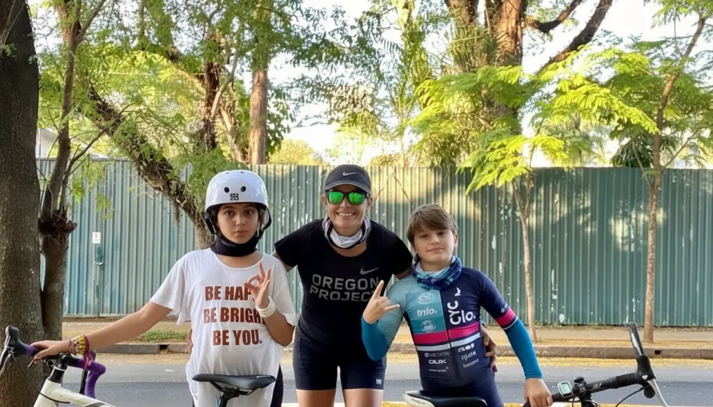 Filhos de Luciana Haddad, usando roupas casuais e bicicletas, em um cenário com árvores e cerca de metal ao fundo.