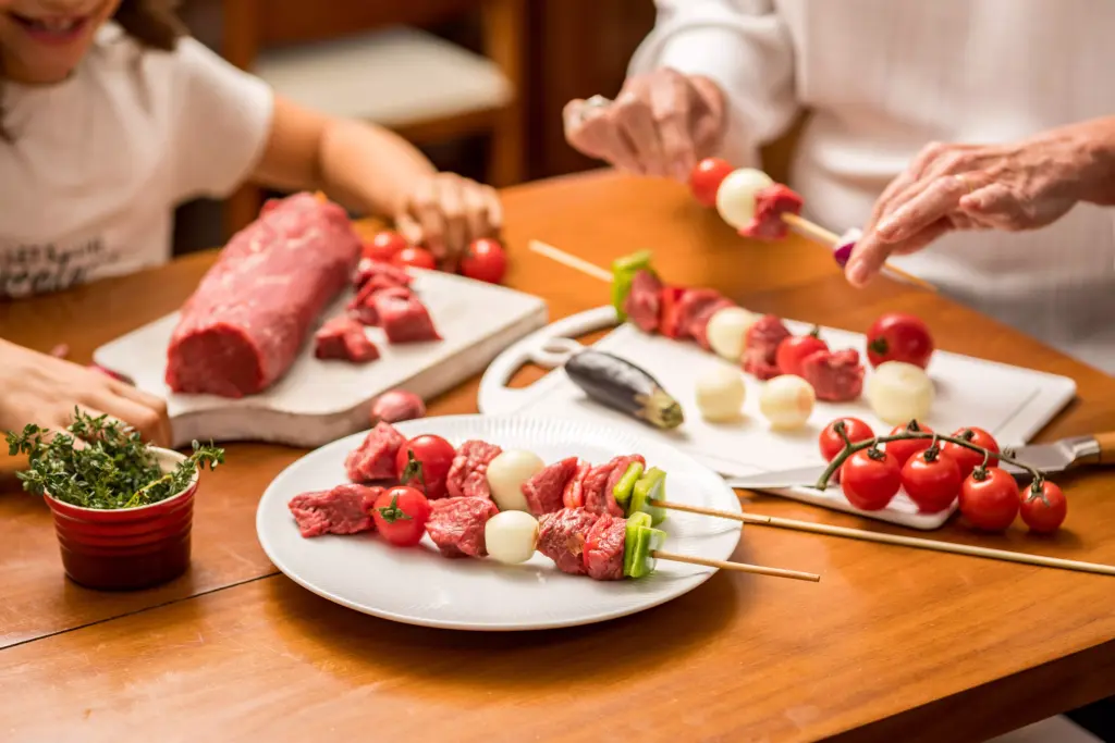 Crianças e idosos preparando espetinhos de carne, destaca a preparação de alimentos saudáveis e atividades culinárias familiares.