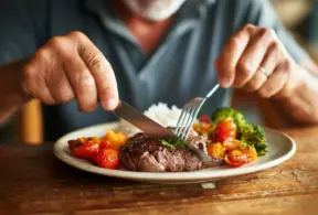 Close das mãos de um homem usando garfo e faca para comer carne vermelha e vegetais, em artigo sobre alimentação e câncer.
