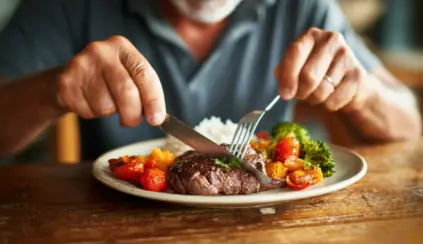 Close das mãos de um homem usando garfo e faca para comer carne vermelha e vegetais, em artigo sobre alimentação e câncer.