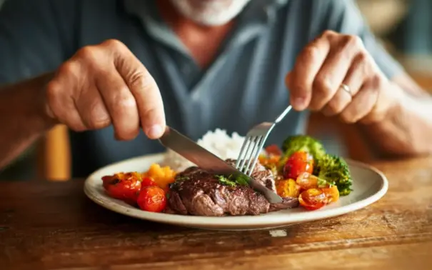 Close das mãos de um homem usando garfo e faca para comer carne vermelha e vegetais, em artigo sobre alimentação e câncer.