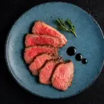 Plato con carne fatiada servida em prato azul com ervas frescas e pingos de molho, em fundo escuro, criando uma experiência do consumidor com apresentação gourmet.