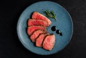 Plato con carne fatiada servida em prato azul com ervas frescas e pingos de molho, em fundo escuro, criando uma experiência do consumidor com apresentação gourmet.