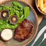 Prato com filé grelhado, folhas verdes e cogumelos fatiados acompanhado de molho branco em tigela e batatas fritas à direita.