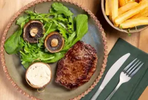 Prato com filé grelhado, folhas verdes e cogumelos fatiados acompanhado de molho branco em tigela e batatas fritas à direita.