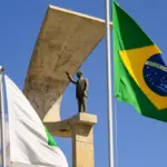 Bandeira do Brasil hasteada ao lado de bandeiras institucionais e de um monumento em pedra com estátua, em um céu azul com nuvens, em ambiente externo