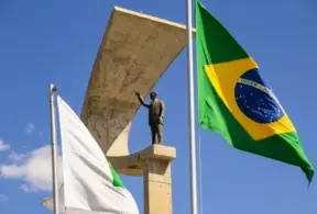Bandeira do Brasil hasteada ao lado de bandeiras institucionais e de um monumento em pedra com estátua, em um céu azul com nuvens, em ambiente externo