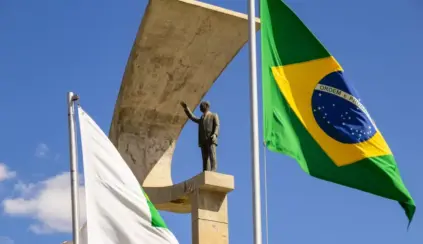 Bandeira do Brasil hasteada ao lado de bandeiras institucionais e de um monumento em pedra com estátua, em um céu azul com nuvens, em ambiente externo