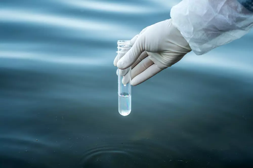 Mão de luvas médicas segurando um tubo de ensaio com líquido transparente, em um fundo de água ou líquido escuro