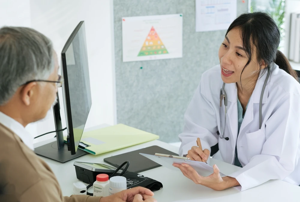 Médica em consultório conversando com um paciente, enquanto analisa documentos e usa um tablet ou computador ao lado de materiais clínicos sobre a mesa.