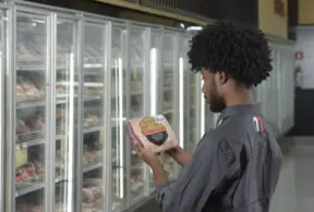 Uma pessoa com cabelos cacheados examina um bife embalado em frente às prateleiras de carne refrigerada, verificando a rotulagem de carnes no Brasil em um supermercado.