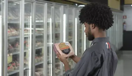 Uma pessoa com cabelos cacheados examina um bife embalado em frente às prateleiras de carne refrigerada, verificando a rotulagem de carnes no Brasil em um supermercado.