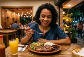 Carne bovina x feijão: Mulher sorrindo em restaurante ao degustar um prato com carne bovina e feijão, usando talheres, com copo de suco e ambiente ao ar livre ao fundo