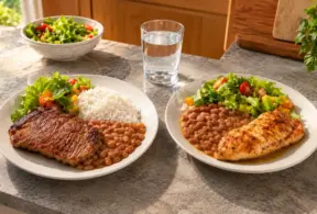 Duas refeições servidas em pratos: à esquerda carne bovina com feijão e arroz, e à direita frango grelhado com feijão, salada e legumes, em mesa com água e folhas verdes