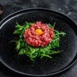 Carne crua em forma de tartare no centro do prato, com gema de ovo e folhas de rúcula