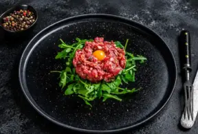 Carne crua em forma de tartare no centro do prato, com gema de ovo e folhas de rúcula