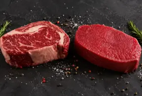 Dois cortes de carne vermelha em destaque sobre uma superfície escura: um bife com marmoreio e outro corte magro, com pimenta-do-reino e ramos de alecrim ao redor — mostrando a diferença entre carne vermelha x carne marmoreio
