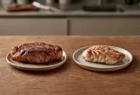 Um bife cozido, rico em nutrientes da carne vermelha, repousa em um prato ao lado de um pedaço de peixe branco cozido em outro prato, ambos sobre uma mesa de madeira com um fundo de cozinha desfocado.