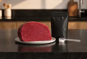 Carne vermelha em fatia em um prato ao lado de um sachê de suplemento de creatina, destacando a diferença entre creatina natural da carne x creatina