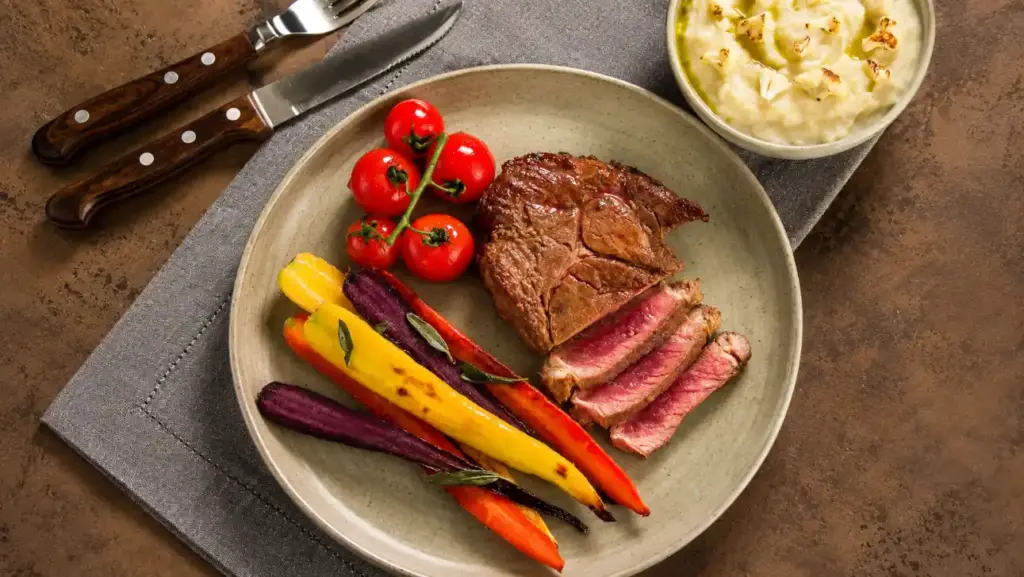 Dieta dos jogadores de futebol em prato com bife grelhado e legumes coloridos como tomate-cereja, cenoura, abobrinha e roxo, além de porção cremosa ao lado
