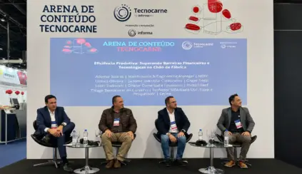 Quatro homens vestidos com trajes profissionais estão sentados no palco, diante de um cenário azul e branco na TecnoCarne. Eles parecem fazer parte de um painel de discussão sobre como superar barreiras à produtividade nas fábricas dentro da cadeia da carne da Tecnocarne.