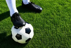 Chuteira de futebol preta sobre a grama artificial com uma bola de futebol no pé, sugerindo preparação antes de entrar em campo e planejamento de pré-treino por modalidade