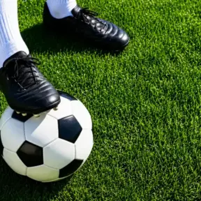 Chuteira de futebol preta sobre a grama artificial com uma bola de futebol no pé, sugerindo preparação antes de entrar em campo e planejamento de pré-treino por modalidade