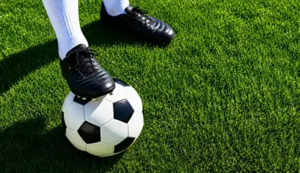 Chuteira de futebol preta sobre a grama artificial com uma bola de futebol no pé, sugerindo preparação antes de entrar em campo e planejamento de pré-treino por modalidade