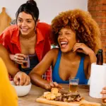 Churrasco em família com pessoas sorrindo e brindando à mesa, ao lado de churrasqueira a lenha de tijolos, com cerveja e petiscos.