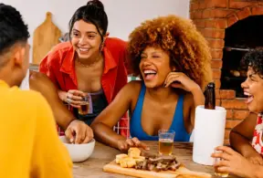 Churrasco em família com pessoas sorrindo e brindando à mesa, ao lado de churrasqueira a lenha de tijolos, com cerveja e petiscos.
