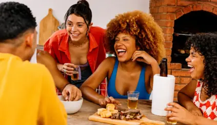 Churrasco em família com pessoas sorrindo e brindando à mesa, ao lado de churrasqueira a lenha de tijolos, com cerveja e petiscos.