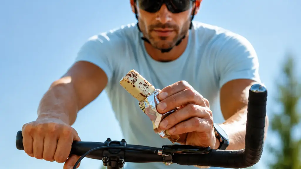 Bicicleta ao ar livre e um homem segurando uma barra de cereal em mãos, sugerindo recuperação muscular pós-treino com reposição energética durante o exercício.