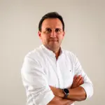 Thiago Bernardino em foto corporativa, usando camisa social branca, com os braços cruzados e expressão confiante diante de fundo liso claro.