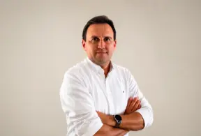 Thiago Bernardino em foto corporativa, usando camisa social branca, com os braços cruzados e expressão confiante diante de fundo liso claro.