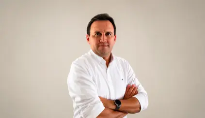 Thiago Bernardino em foto corporativa, usando camisa social branca, com os braços cruzados e expressão confiante diante de fundo liso claro.