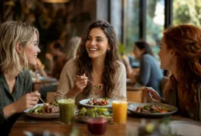 Três pessoas sorrindo e conversando durante um almoço em restaurante, com pratos e sucos na mesa, em clima de socialização e consumo consciente, simbolizando indulgência limpa e novo hábito de consumo na Europa.
